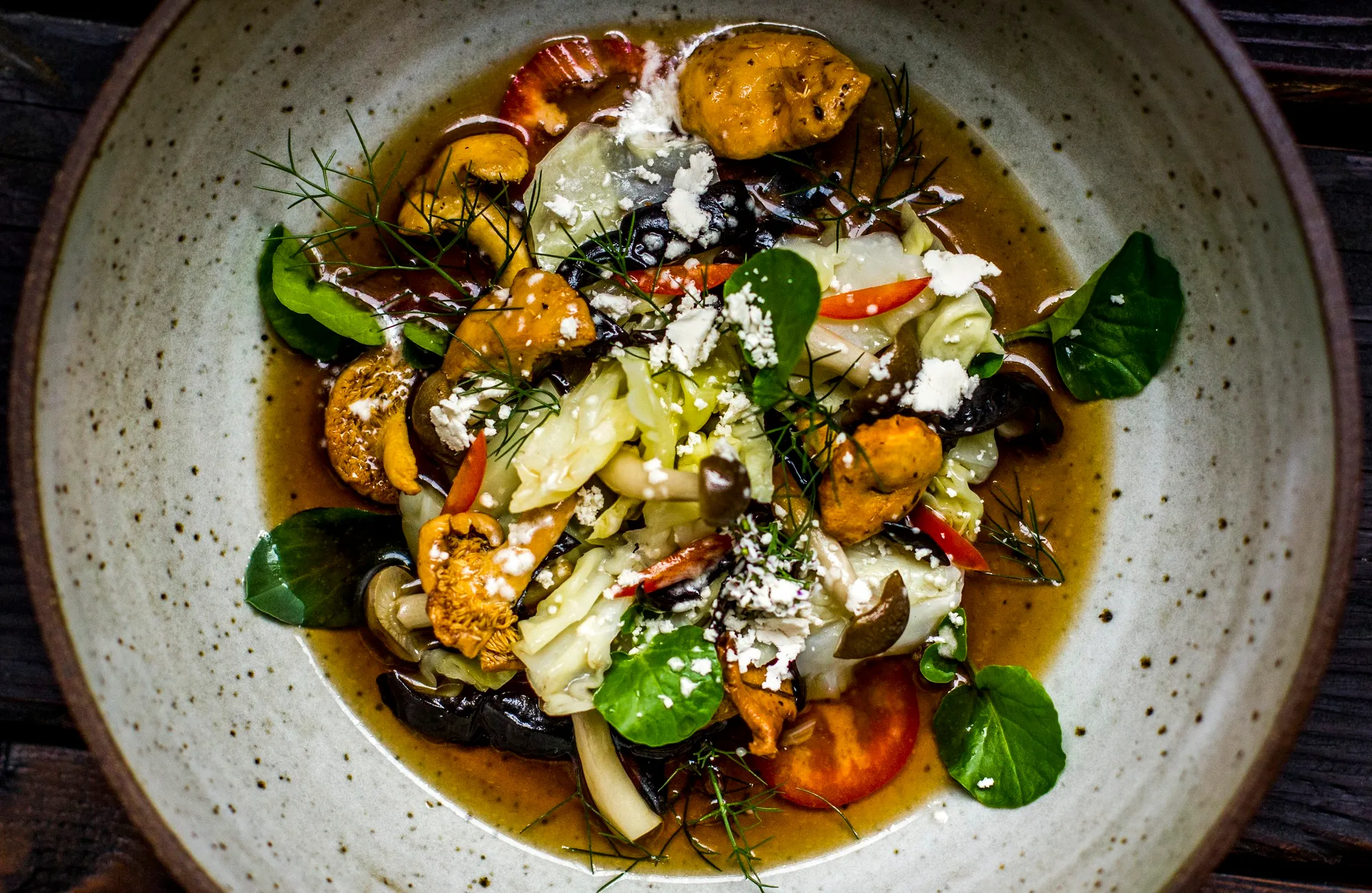 An overhead bowl of chanterelles, watercress, cabbage and tomato in a golden broth, scattered with crumbled white cheese and dill fronds