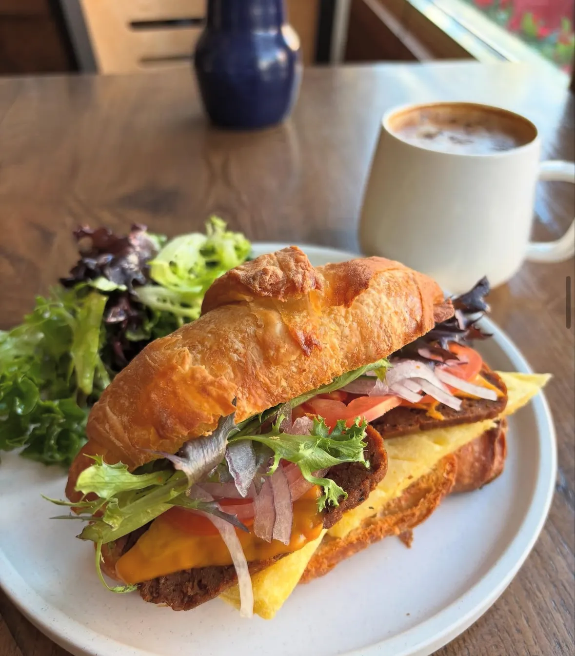 A croissant sandwich filled with a breaded cutlet, melted cheese, tomato, red onion and lettuce, served with a side salad and a cup of coffee