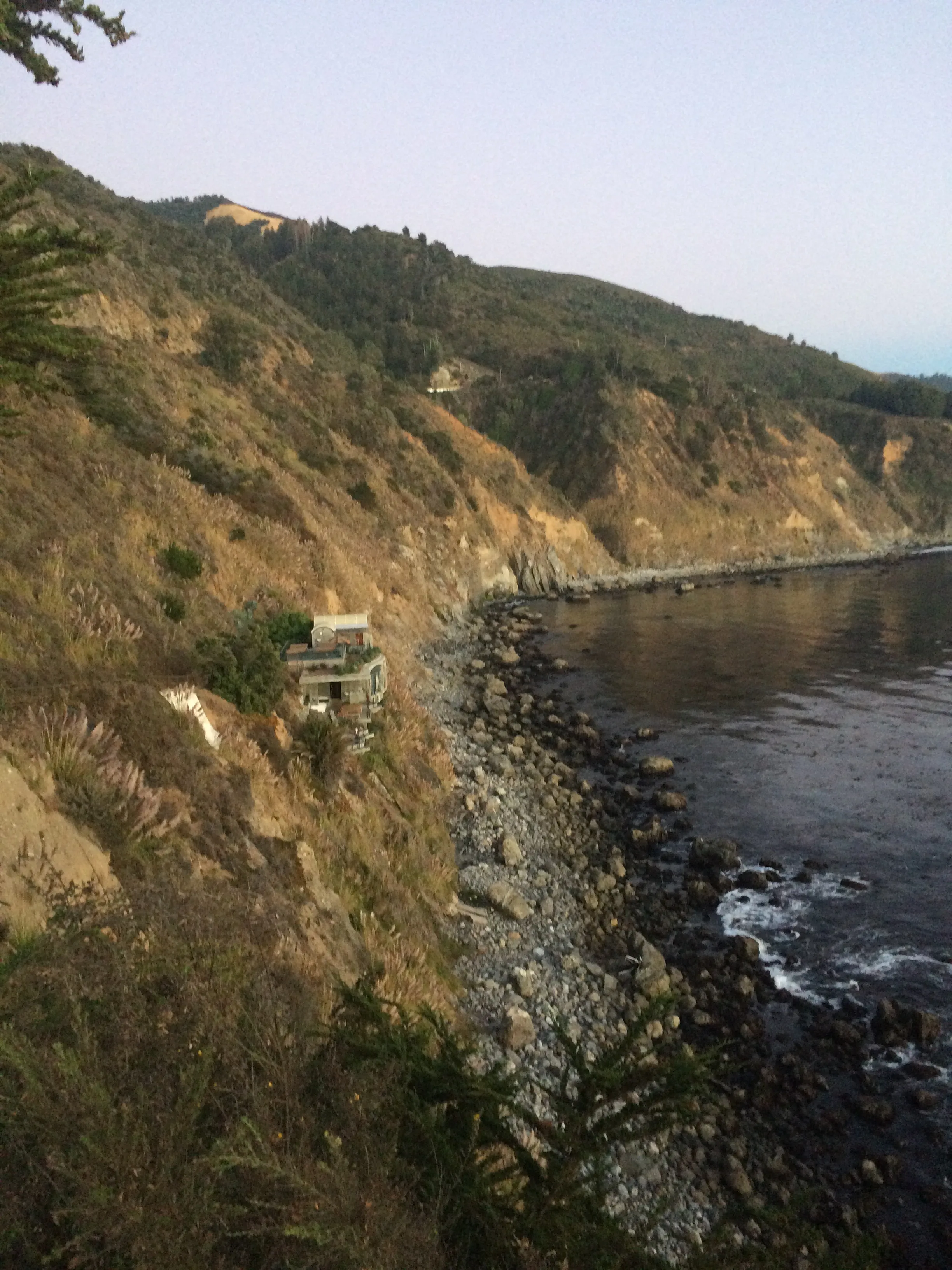 The Big Sur coastline at dusk from above Esalen — a low cliffside building among cypress, a rocky shore, and the Pacific running out to the horizon
